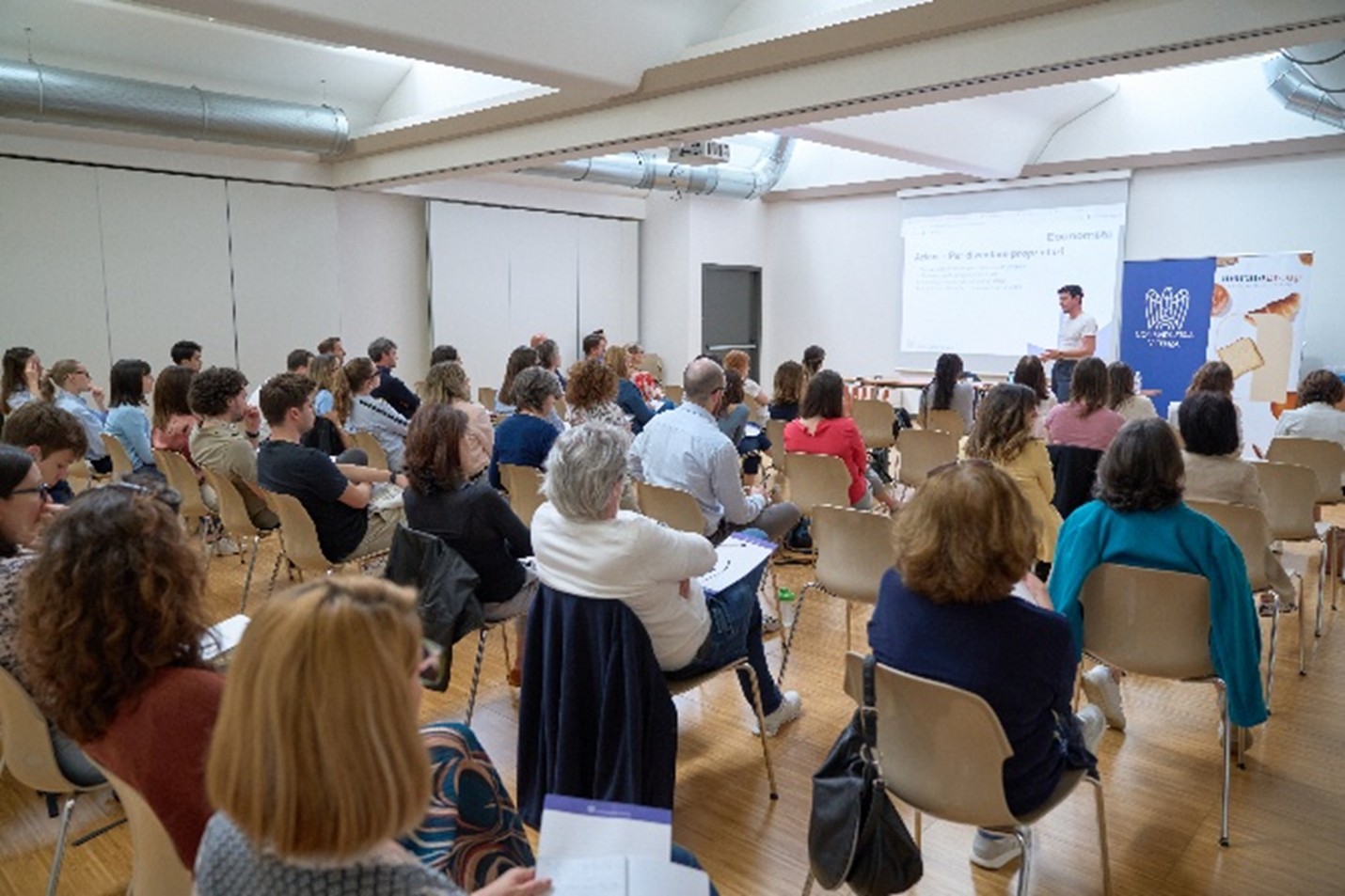 A packed hall during a seminar with participants listening to a speaker.